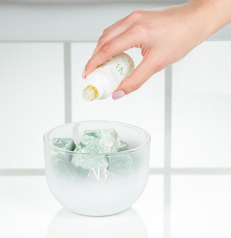 Hand pouring a white bottle into a clear bowl with green contents on a light background