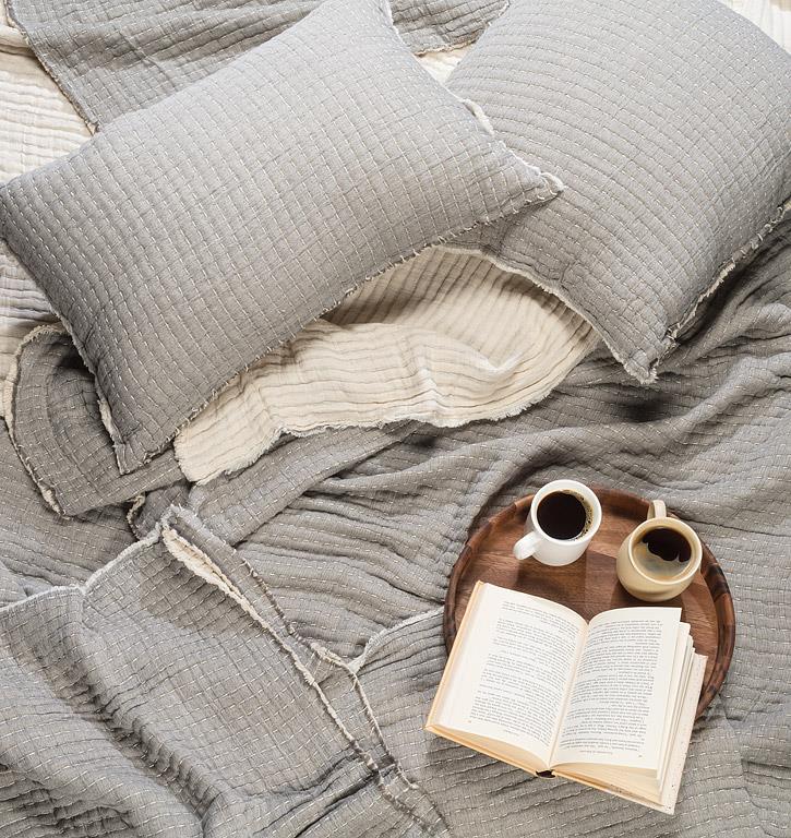 Gray textured pillows and blanket with a book and coffee on a wooden tray.