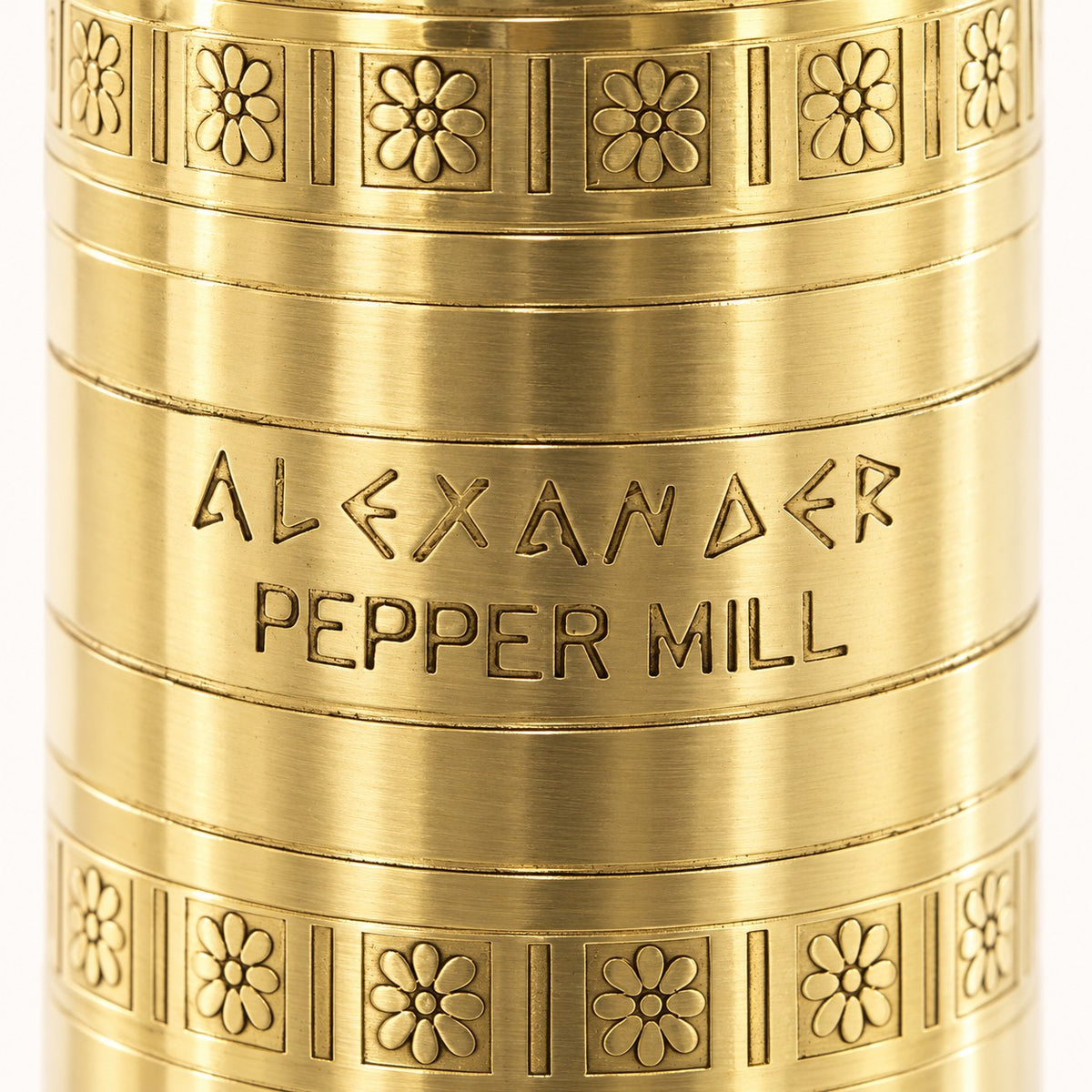 Close-up of a brass-colored metal pepper mill body with engraved text reading 'ALEXANDER PEPPER MILL' and decorative floral patterns around the top and bottom edges.