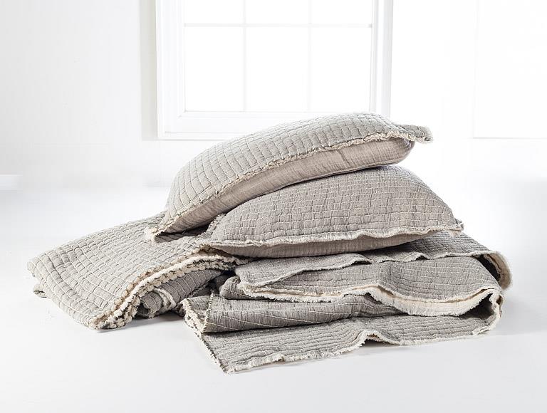 Stack of textured gray pillows and blanket on a white surface with a window in the background.