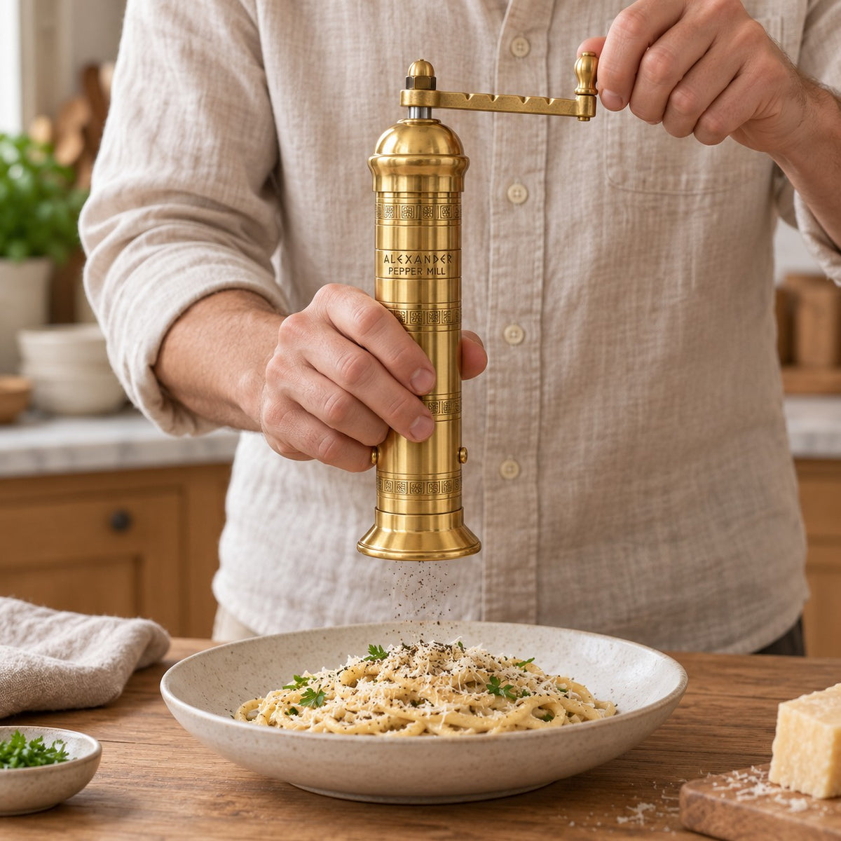 A person in a beige linen shirt grinds black pepper from a tall brass pepper mill over a bowl of pasta on a wooden table in a warm kitchen setting.