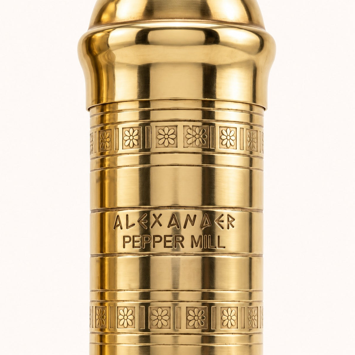 Close-up of a polished gold metal pepper mill body with engraved floral bands and the text 'ALEXANDER PEPPER MILL' on a light neutral background.