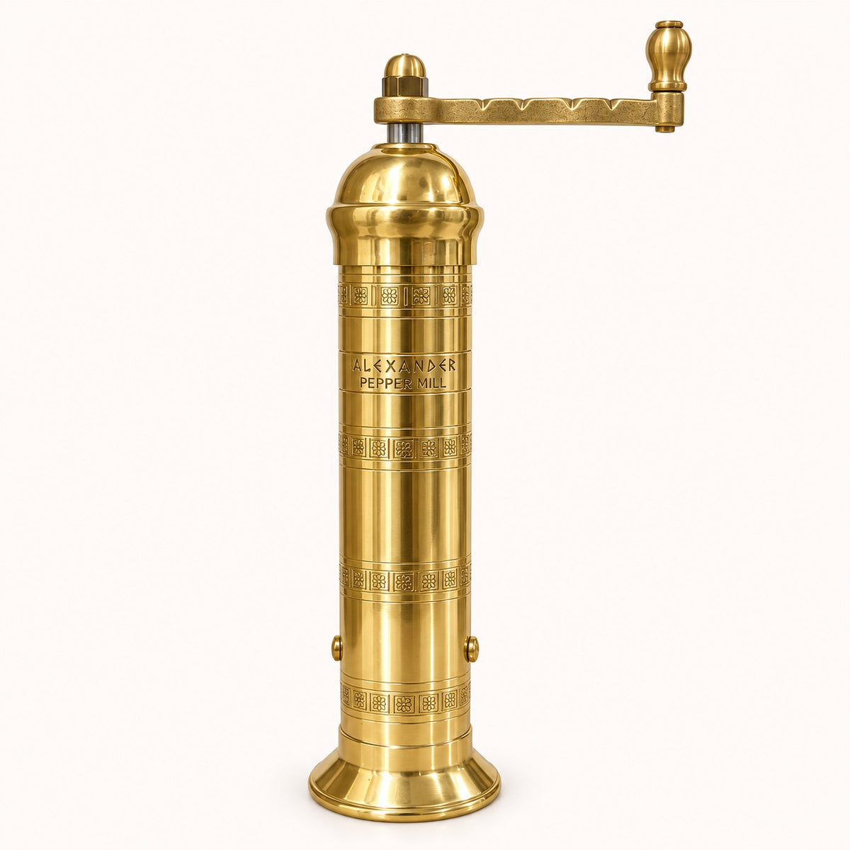 A vintage-style brass pepper mill with a cylindrical body, engraved decorative bands, and a horizontal crank handle, shown on a plain light background. The body is engraved with the text 'ALEXANDER PEPPER MILL'.