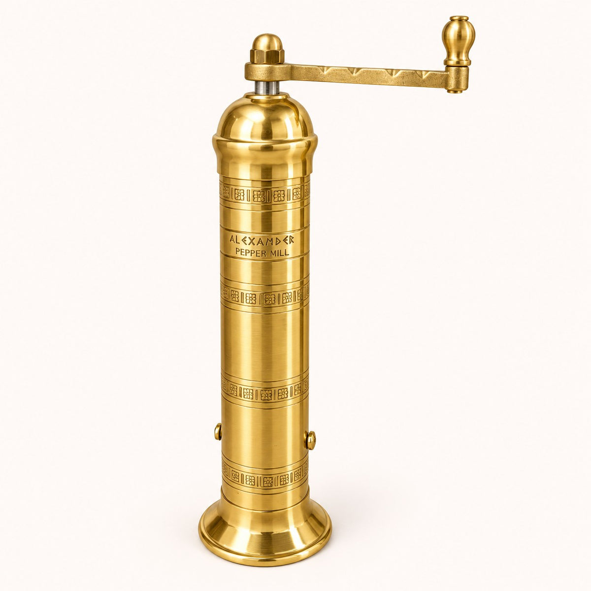 A vintage brass pepper mill with a cylindrical body, decorative Greek key engravings, and a horizontal handle with a ball finial, shown on a light cream background.