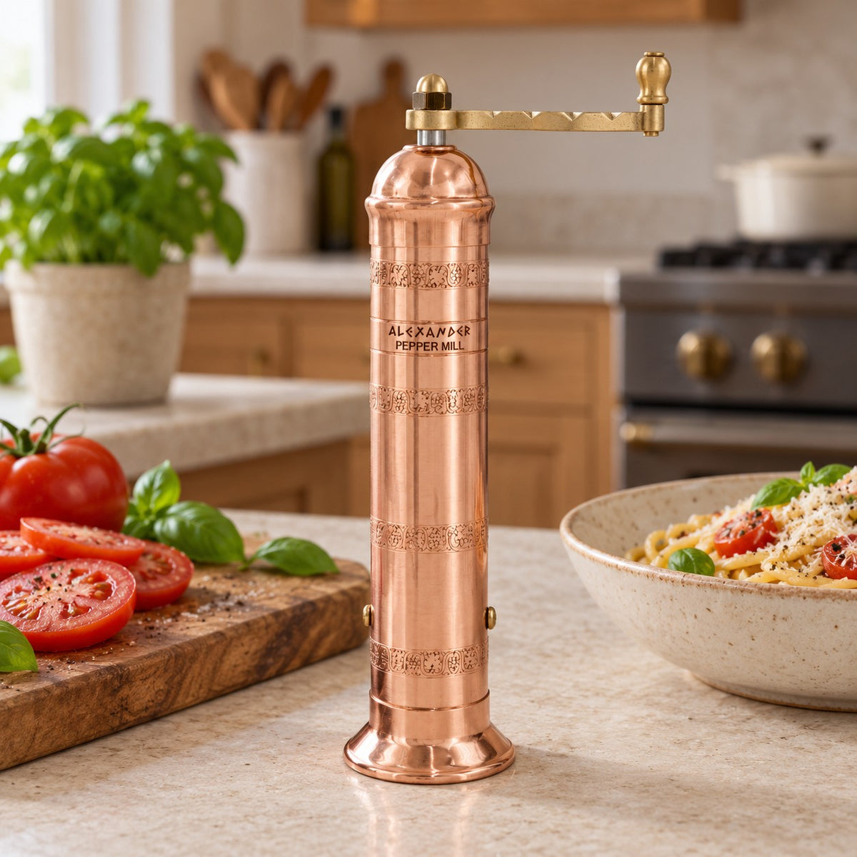 A copper manual pepper mill with a brass crank handle stands on a kitchen counter, surrounded by fresh tomatoes, basil, and a bowl of pasta in a warm, blurred kitchen setting.