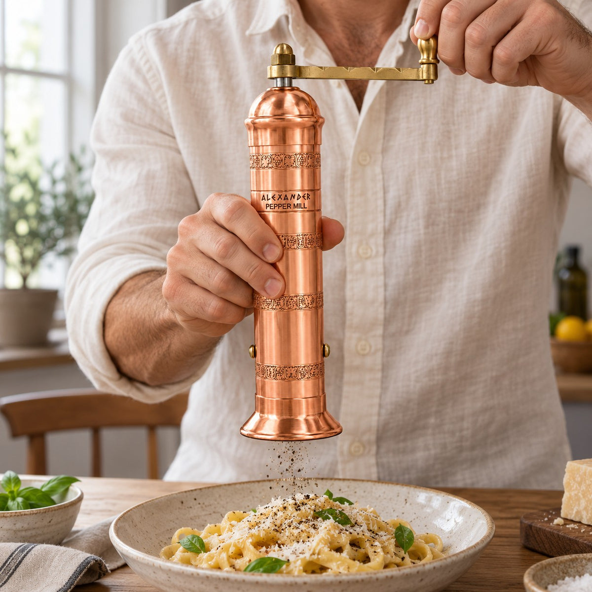 A man in a white linen shirt uses a copper and brass manual pepper mill to grind pepper over a bowl of pasta with basil in a bright kitchen setting.